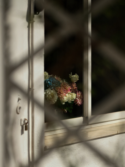Durch einen Maschendrahtzaun der Blick auf ein geöffnetes Fenster, die Sonne scheint auf einen Plastikblumenstrauß.
