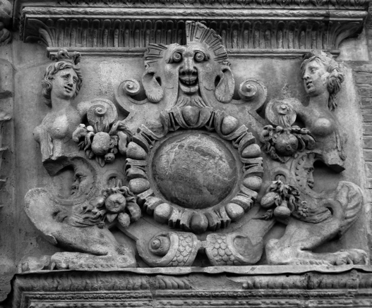 Close up photograph in black and white of a detail of the Renaissance portal of a hotel, showing a fantastical character, with broad smile, round eyes, goat ears and beard, and sun like hair. On each side, two female busts seem to be looking and laughing at this sight. A globe, maybe the sun, fruits, flowers, crescent moons and lion feet complete the decor of this trio.