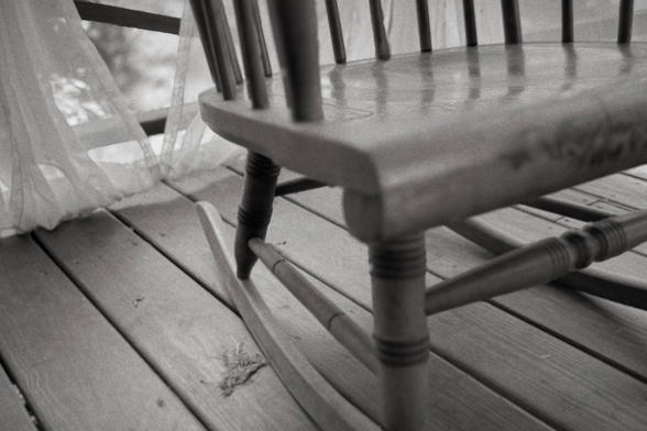 This photograph shows the lower portion of a wooden rocking chair on a porch, viewed at a diagonal angle. Sheer curtains billow gently on the left side, and the wooden floorboards run diagonally beneath the chair’s curved rockers. The scene is softly lit and slightly out of focus, giving it a quiet, dreamlike atmosphere.