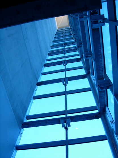 Color photo looking up through an open shaft that is defined by a smooth concrete wall (left) and two walls of metal framed blue glass (center and right)