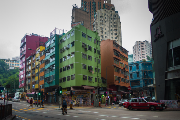 An urban street scene with colorful buildings and a red taxi in the foreground. Pedestrians are crossing the street, and there's a mix of modern and older architecture.