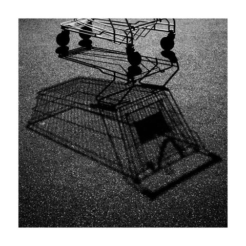 A square black-and-white photograph framed with a white border, showing the lower part of a metal shopping cart casting a sharp, distorted shadow onto coarse asphalt. The high-contrast lighting emphasizes the cart’s wireframe structure and the textured surface of the ground. The shadow appears almost like a full 3D duplicate, shifted and stretched across the pavement, creating a surreal doubling effect. Only the wheels and base of the actual cart are visible at the top edge of the frame. The image is minimal, graphic, and abstract in character.