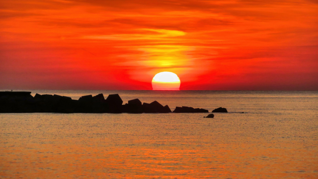 La imatge mostra una posta de sol impressionant sobre el mar. El cel està pintat en tons taronges i vermells vibrants, mentre que el sol es troba just a sobre de la línia de l'horitzó, creant un efecte càlid i lluminós. En primer pla, es poden veure roques fosques que contrasten amb els colors brillants del cel i l'aigua, que reflecteixen la intensitat de la posta de sol. L'escena transmet una sensació de pau i tranquil·litat, típica d'un moment màgic al final del dia.