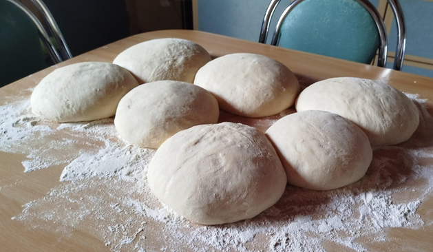 Des boules de pâtes à pain sont disposées sur la majeure partie de l'image.
Il y en a 7. Elles sont posées sur une table en bois brun clair. Il y a de la farine tout autour et sur le bas de l'image.
À l'arrière plan, courbes métalliques et vert  de dossiers de chaises 