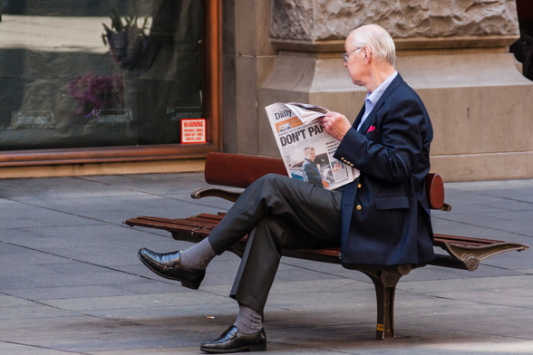 A conservative looking, elegantly dressed older man is sitting on a bench reading a newspaper. On the front page, you can only see the first word of the newspaper title, Daily, at the top. Below, in very large uppercase letters, there's a heading that says, DON'T PANIC. This is obvious, even though the second word is partially obscured at the end.