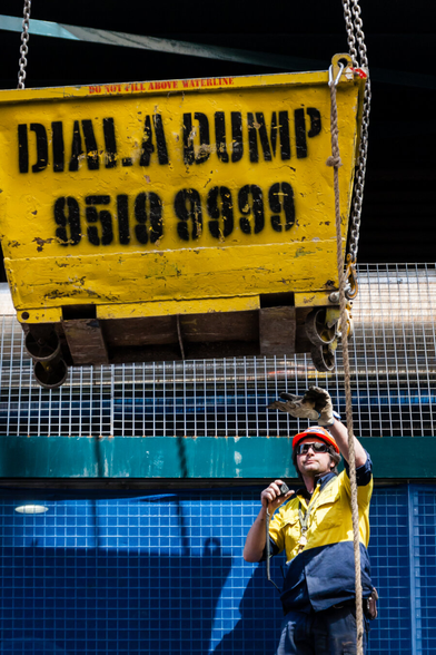 At the lower right, a construction worker with a hard hat and sunglasses is looking up, seemingly to signal for the movement of the very large container that is suspended by chains here. On it, are the words, Dial a Dump, and below that, there is a phone number. In the background is a steel cross beam and grid like metal wires, which are part of the construction site.