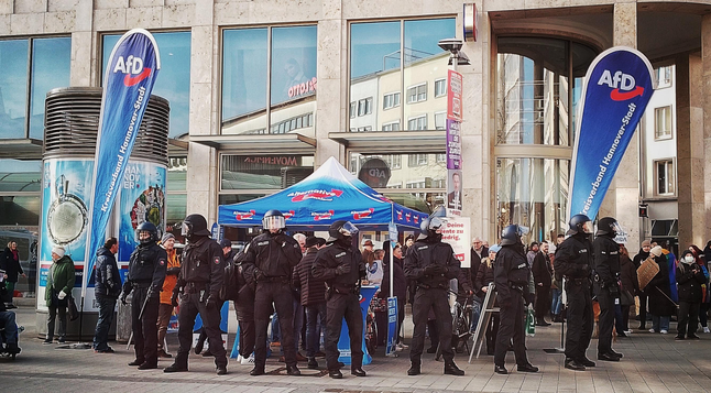 Alteres Foto, Hannover, Kröpcke. Fußgängerzone. Schaufenster. Davor: blaues Zelt, Flaggen, Aufsteller, blau mit AfD-Logos. Davor: eine Reihe Polizisten, Vollschutz, Helme. Verhindern ein Näherkommen der demokratischen Demonstranten — die die Rechtsextremen gerne einmal inhaltlich stellen würden.