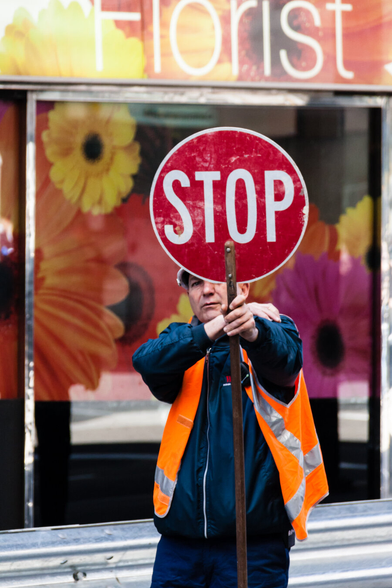 A road worker in an orange safety jacket holds up a red and white stop sign for traffic. His face is partially obscured by the sign, but the stare from the one eye visible is quite intense. Behind him is a colourful flower pattern on a shop front, and at the top on the wall is the word, Florist.