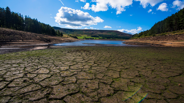 a half empty reservoir with a bottom that is cracked on a bright blue sky with clouds and hills with trees around it.  