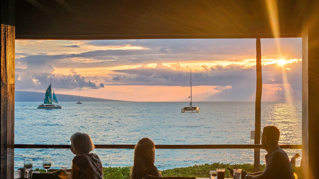 A warm sunset view from an oceanfront restaurant, framed by wooden beams and soft golden sunlight streaming in from the upper right. Three people are seated at a table in the foreground, silhouetted against the glowing horizon. Two sailboats drift peacefully on the ocean, one with teal and white sails on the left and a catamaran centered in the distance. The water reflects the golden hues of the sky, and lush greenery lines the edge of the patio. Glasses of wine and cocktails sit on the table, enhancing the tranquil, tropical atmosphere.