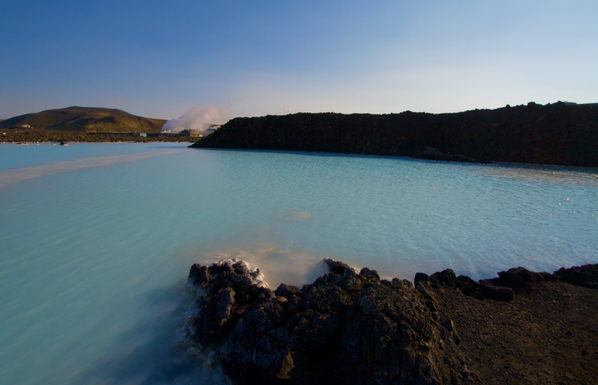 an expanse of shades of blue can be seen in this photo of Iceland's blue lagooon. The lagoon stretches from the bottom of the photo to the opposite shore, near the middle, where it meets low mountains and dark hills. some of the dark ground juts into the lower right, and it all sits under a brilliant blue sky that goes near white at the right edge. a tuft of steam is seen near the center and a line of grey shoots thru the upper left