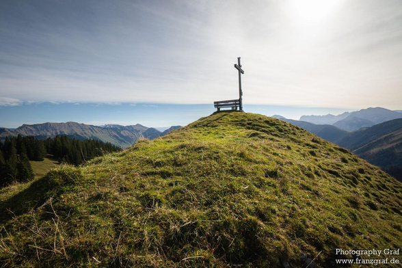 A serene scene captured on a hill, showcasing a wooden bench with a simple cross on top. The hill overlooks a vast landscape with a clear blue sky and fluffy white clouds. The setting is peaceful and tranquil, surrounded by mountains and greenery. The dominant colors in the image are white and a warm brown accent. The image exudes a sense of spirituality and contemplation, inviting viewers to sit and reflect on the beauty of nature. This picturesque hill station offers a perfect spot for relaxation and connecting with the natural world.