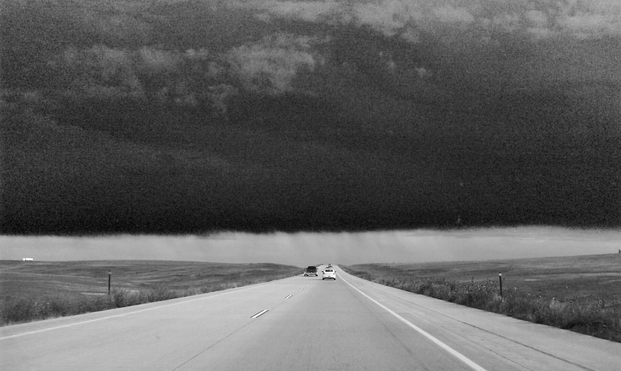 A black and white image of a long, straight highway stretching into the distance. Dark, ominous storm clouds loom overhead, creating a stark contrast with the light-colored road. Two cars can be seen driving ahead on the highway, which is surrounded by flat, grassy plains. The scene captures the dramatic approach of a severe weather system over an open landscape.