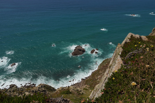 Photo of the rocky cliffs of Portugal. In the foreground there is some green on the grey, rough rocks that fall off sharply into the blue-greenish ocean below. The waves batter a rocky outcrop.