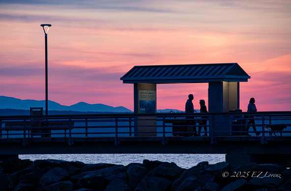 People silhouetted against the sunset sky in pink glow walking on a fishing pier at Edmonds, Washington with the partial view of the Olympic mountains in the background. 