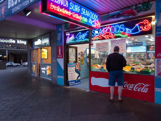 The back view of a man in shorts can be seen looking into the window of a fish and chip shop. It is night time and everything is lit up very brightly. The flourescent signs outside and above the shopfront say, Bondi Surf Seafoods. On the left, another shop at the street corner is called, Noodle King, but this is not the primary focus of the image.
