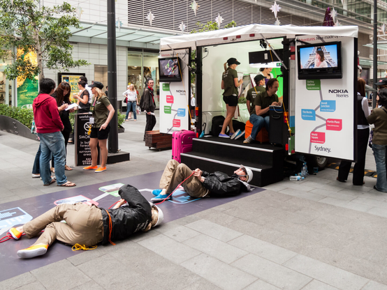 In an urban pedestrian area, there's a mobile studio with two steps leading up to it. There is advertising for Nokia on the outside. Inside the studio, four people can be seen, and one is at a computer keyboard. Lying on the ground outside, two men with hard hats are holding onto a rope, and look like they are scaling a building or something. The scene is puzzling.