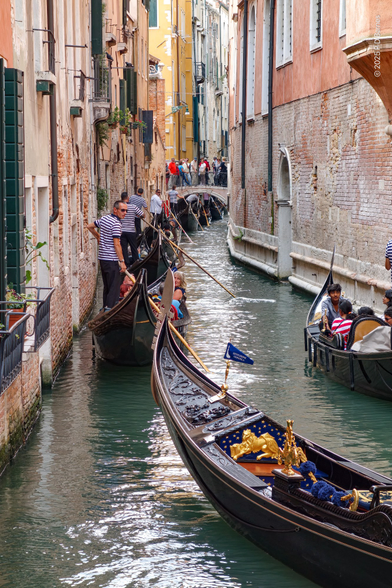 A narrow Venetian canal is completely full of stem-to-stern gondolas waiting to progress under a bridge and down the canal. 