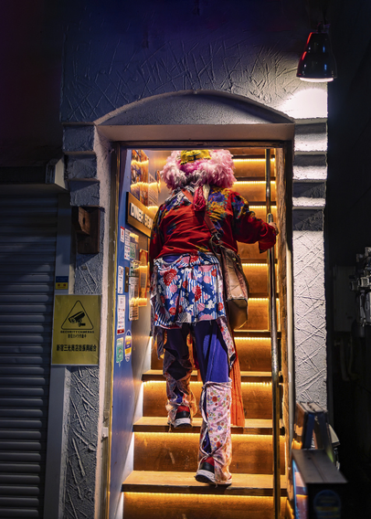 A man dressed in a colourful fancy costume is entering a narrow entrance and walking up straight narrow illuminated stairs in a dark alley in Shinjuku, Tokyo.