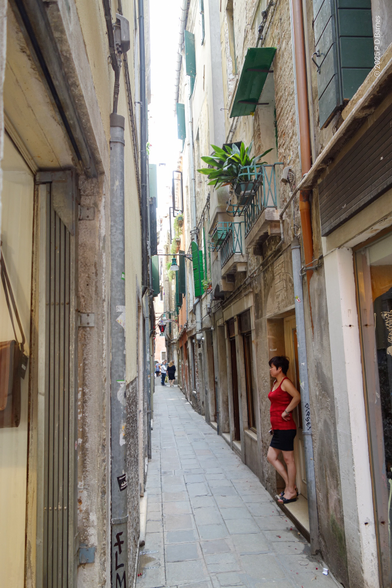 A narrow street in Venice. A woman is standing in a doorway, hand on hip, waiting for something or someone.
