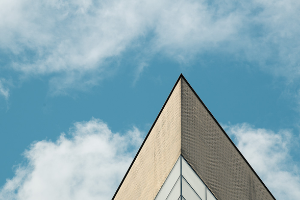 Die spitz zulaufende Ecke eines Bürogebäudes ragt in den blauen Himmel, der mit ein paar Wolken versetzt ist. Das Gebäude wird von der Sonne bestrahlt, so dass sich links die helle und rechts die schattige Seite befindet. Das Bild wirkt insgesamt recht abstrakt und geometrisch.