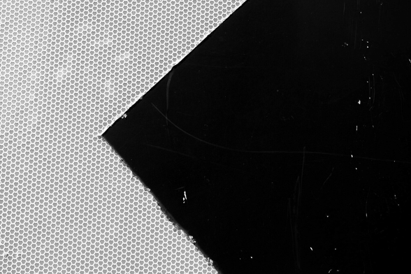 Black and white, high contrast photo of a close-cropped section of a large black and white 'bear left' road sign. On the right, the black background, lightly pock-marked; on the left, part of the interior of a white chevron, which is actually made up of a repeating pattern of roughly hexagonal reflective shapes.