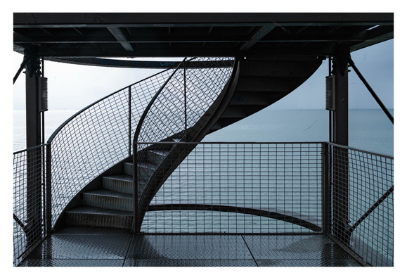 Foto im Querformat. Blick auf eine Wendeltreppe an einem See. Unten ist eine Plattform mit quadratischen Steinfliesen. Auf allen Seiten ist ein Geländer aus Metall mit rechteckigem Gitter. Links starten die Wendeltreppe und geht nach rechts oben. Die Treppe hat ebenfalls ein Geländer mit Gitter. Oben im Bild ist die nächste Plattform, die Unterseite ist ein Gitter aus Stahl. Im Hintergrund ist ein See. Es ist ein regnerischer Tag, alles ist bläulich grau oder schwarz.