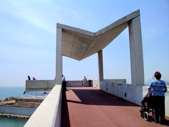 The image depicts a modern architectural structure with a concrete framework, featuring a distinctive V-shaped roof supported by two vertical columns. The structure is part of a pathway that extends over a body of water with a clear blue sky in the background. The pathway is paved with a reddish-brown surface, and there are people visible. The architecture is minimalist, with clean lines and a focus on geometric shapes. The surrounding area includes a low concrete wall and some construction materials, suggesting ongoing development. The overall scene is bright and open, with a sense of openness and modern design.

Provided by @altbot