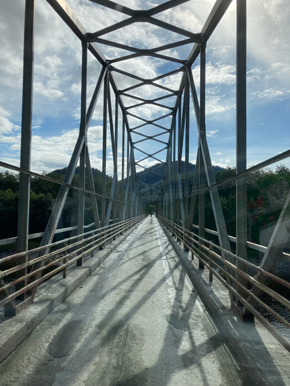 Zu sehen ist eine schmale Brücke. Die Brückenkonstruktion besteht aus vielen Metallstreben. Im Hintergrund sind Berge und Wald zu erahnen.