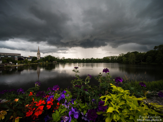 Photographie en couleur présentant un ciel d'orage au-dessus d'un étang, près d'une église, et avec une jardinière de fleurs au premier plan. L'ambiance générale est relativement sombre avec de gros nuages gris très foncé, alors que certaines zones bénéficient encore de la lumière solaire comme la jardinière au premier plan et le clocher de l'église, à gauche, qui se reflète parfaitement sur la surface de l'étang, et plus loin à l'horizon où il semble qu'on devine une averse sous un nuage. À droite de la photo, l'étang est bordé par un petit bosquet. Des fleurs de la jardinière sont rouges, violettes avec des feuilles vertes et jaunes.