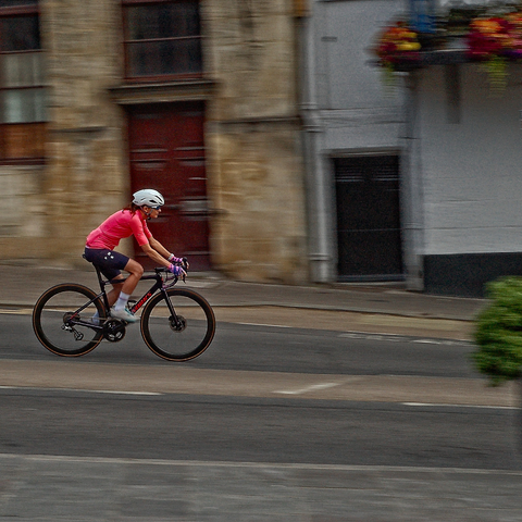 a cyclist weaing a white helmet, salmon pink top and black shorts and riding a black racing bike zips down a hill, past buildings of cotswold stone.  The cyclist is in focus whilst all around is blurred.  