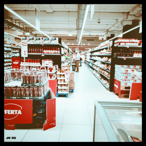 Farbfoto in etwas blassen Farben mit Rotbetonung, aufgenommen in einem italienischen Supermarkt. Rechts vorn eine Gefriertruhe, links vorn eine Angebotsinsel mit Tomatensoßeflaschen, Weiter entfernt ein Gang mit Regalen und Waren links und rechts. Oben die Decke mit Leitungen und Leuchtmitteln. Im Gang befinden sich zwei Einkaufende.