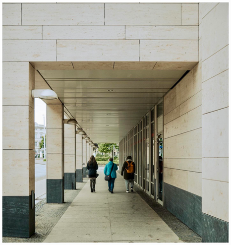 Ein überdachter Gehweg mit drei Personen, die sich von der Kamera entfernen. Die Architektur verfügt über helle Steinmauern und eine glatte, gepflegte Oberfläche. Es werden Elemente wie Säulen, Deckenbeleuchtung und Schaufenster aus Glas angezeigt.