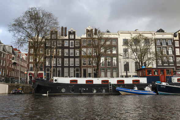 View from a boat on a canal in a city. We're looking at the metre-high stone bank where some boats are docked in front of four and five storey town houses. The largest of the boats looks to have been a former canal cargo carrier now converted to living accommodation on the water and it has a motorised dinghy tied to it. A few of the buildings behind the boats have small stepped gables. The sky is overcast but there's a warmth in some of the light hitting the scene that suggests early evening with the sun going down somewhere behind us.