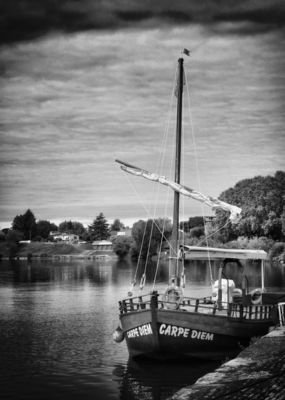 Ein Schwarz-Weiß-Bild eines Bootes mit dem Namen "Carpe Diem", das an einem Flussufer angedockt ist. Das Boot hat einen hohen Mast und ein Segel, im Hintergrund sind Bäume und Häuser zu sehen, die sich im ruhigen Wasser spiegeln.