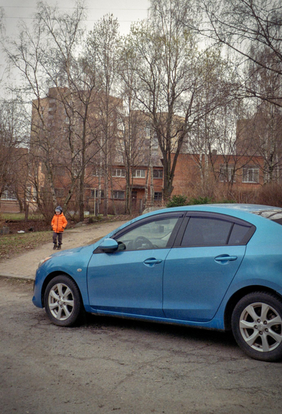 There is a blue car in the foreground, and a child in an orange jacket is walking next to it. Beyond them is a park and houses.