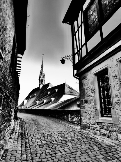 Auf dieser Schwarz-Weiß-Aufnahme erkennen wir eine mittelalterliche Kopfsteinpflastergasse, links und rechts von Häusern gesäumt, weiter vorne links unterhalb einer Mauer ein gotischer Hallen- Kirchbau mit einem Dachreiter. Es handelt sich hierbei um eine Straße in der Alten Hansestadt Warburg in Westfalen.