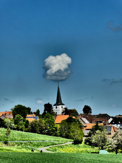 Wir schauen auf die Kirche von Ottenstein, dem Hauptort der Ottensteiner Hochebene im Weserbergland.  Der Himmel ist fast blau und eine große Wolke scheint auf der Spitze des Kirchturms zu balancieren, ihre Unterseite ist graublau, die Oberseite weiß.