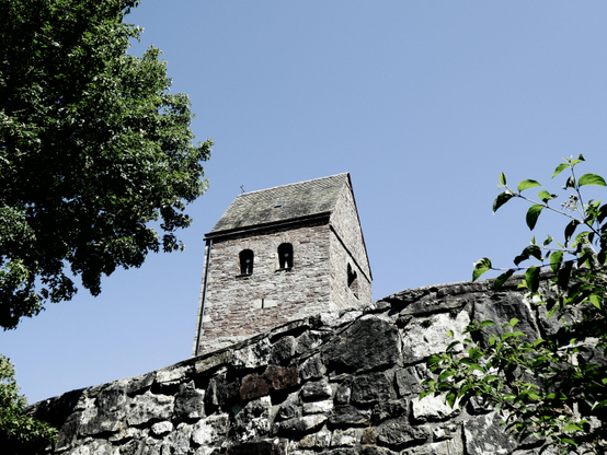 Hinter einer Bruchsteinmauer ist der sandsteinerne Turm einer romanischen Kirche zu erkennen, links im Bild ein Baumwipfel. Wir schauen dabei von unten nach oben.