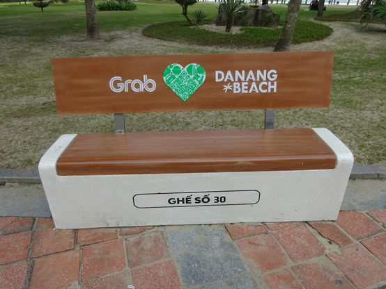 Parkbank mit Holzlehne und Sitzfläche, Aufschrift 'Grab', grünes Herz mit QR-Code, 'Danang Beach' und 'GHẾ SỐ  auf Betonbasis