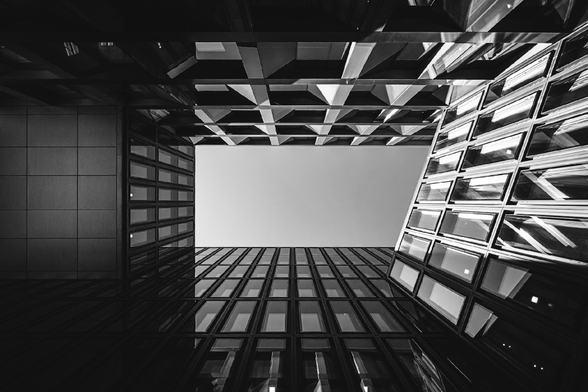 Black and white photo looking up through the narrow opening between modern glass buildings, with geometric window patterns and reflections creating a sharp, angular frame around a pale, cloudless sky.
