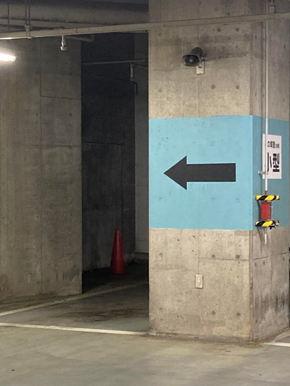 A parking garage corner features a gray concrete pillar with a light blue section, an arrow pointing left, and a red traffic cone visible in the background. A security camera is mounted on the pillar.