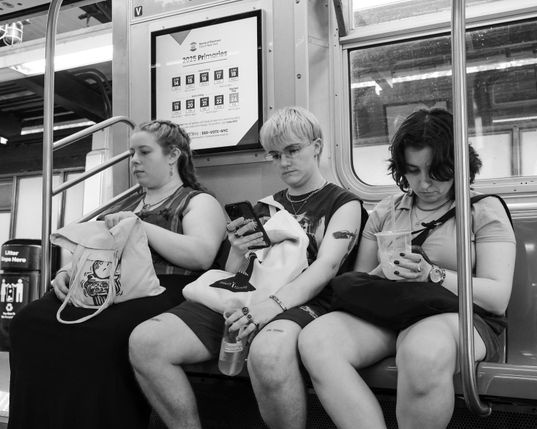 Black and white photo of three young people sitting next to each other in a subway car. Tow are looking at their phones, one is looking into their handbag.