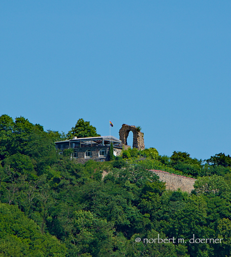 Steinerner Mauerbogen auf einer mit grünen Bäumen und links einem Ausflugslokal umgebenen Hügelkuppe, mit strahlend blauem Himmel