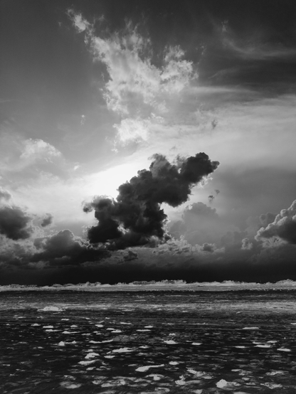 Dramatic black and white photo of the setting sun behind some storm clouds over a sandy beach.