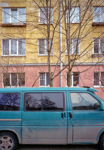 A close-up of a blue minibus, followed by a multi-storey house, the bottom of which is pink and the top is ochre.