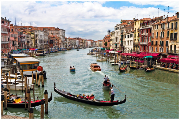 A scenic view of the Grand Canal in Venice, featuring traditional gondolas and wooden boats navigating the water. Colorful buildings line the canal, with outdoor dining areas under awnings and a partly cloudy sky overhead.