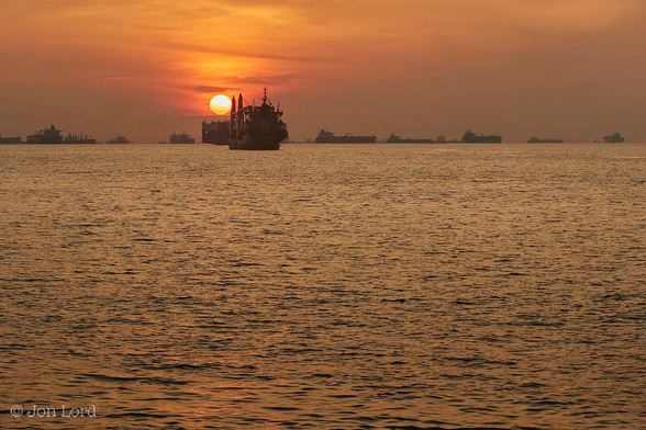 This Is A Colour Seascape Photo of a tropical sunrise and a long row of anchored merchant ship In Landscape Format. The Entire Image Is Tinted With A Golden Glow From A Just Risen Tropical Sun (Only A Few Degrees Above The Horizon). A Calm Sea Covers The Lower Three-Quarters Of The Frame With A Crisp Horizon Dividing The Ocean From The Sky. The Water Has Numerous Ripples Disturbing The Surface But Is Essentially Calm. Stretching Across The Horizon Is An Anchorage With A Great Many (Twenty-One In View) Cargo Ships At Anchor. The More Distant Vessels Are At Right Angles To The Camera Though The Two Closest, Above And To The Left Of Centre, Are A Medium Sized Container Ship And Car Carrier Are Nearly Parallel To The Lens. Just Above These Two Ships Is The Golden Rising Sun That Has Cast A Glow Across A Thin Band Of High Level Cirrus Clouds. 
The Location: The Singapore Straits, South China Sea (2017).
