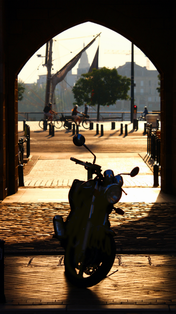 Photo en contre jour, au lever du soleil, la lumière ressort dorée.
Une moto est en premier plan, dans l'encadrement d'une arche de pierre qui mène aux quais.
On aperçoit l'autre rive, en dernier plan, des cyclistes sur la bande cyclable qui longe le quai.
Et le mat d'un des voiliers présents pour le festival du vin.