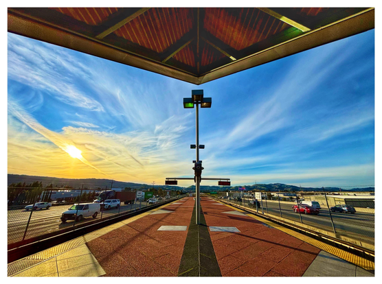 Landscape-orientation photo of the open-air station platform for the BART train in Dublin, California. The roof overhang and the platform surface both form visual triangles at the top and bottom of the image, as they recede towards a signal post standing at the center. Other symmetrical geometric patterns are formed by the beams and rows of parallel lines on the underside of the overhang, as well as the variously configured tiles on the platform, which are predominantly in yellow and reddish brown. Freeway traffic flows on either side of the platform. A line of low mountains sits at the horizon. The vivid blue sky is streaked with white cirrus clouds and washed in golden light at left frame by the afternoon sun.